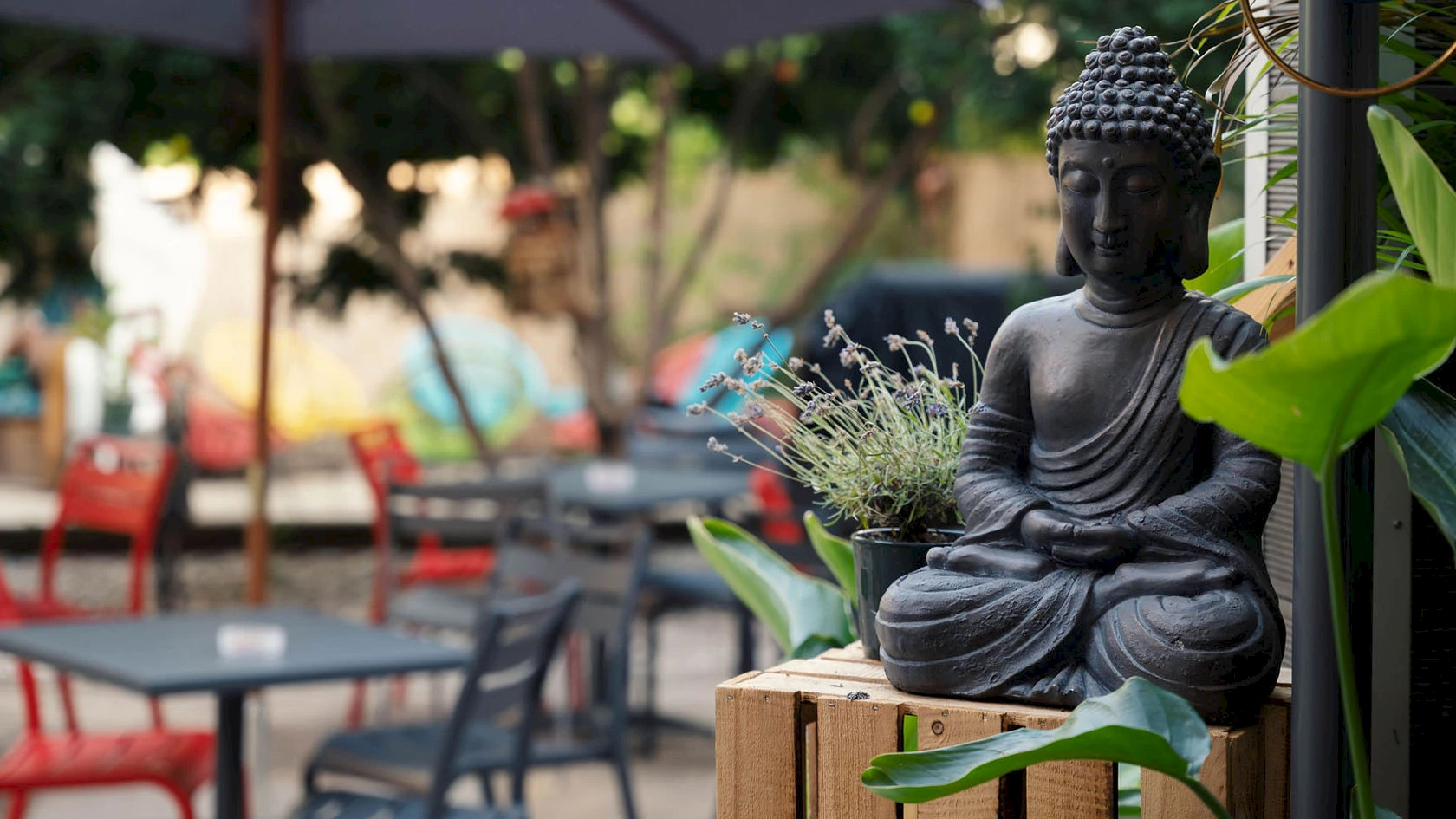 Im Außenbereich stehen Tische, Stühle und Sonnenschirme; auf einer Holzkiste sitzt eine Buddha-Figur.